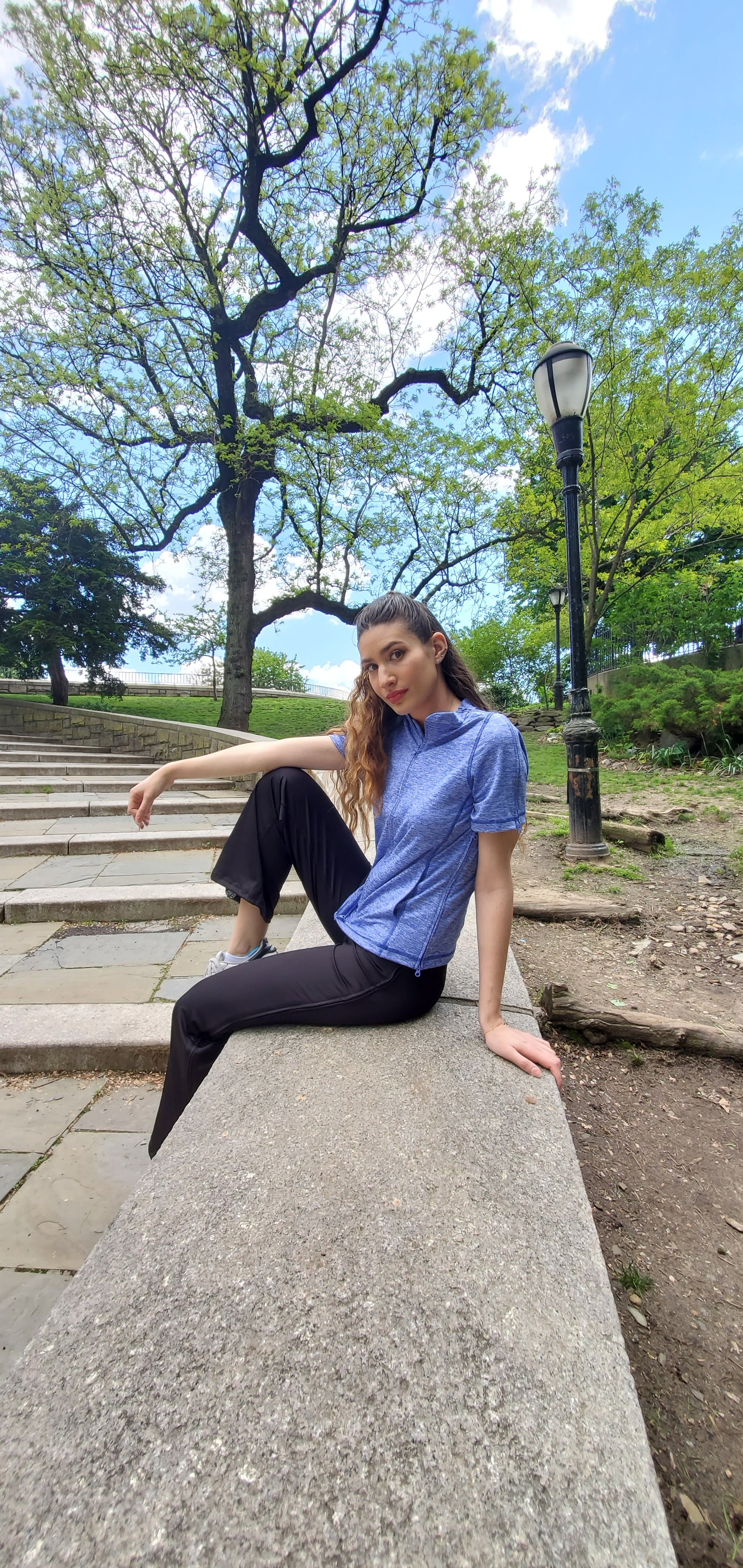 Girl with brown wavy hair sitting on a stone wall by steps in the park, wearing a blue Reboundwear top and black Reboundwear pants.